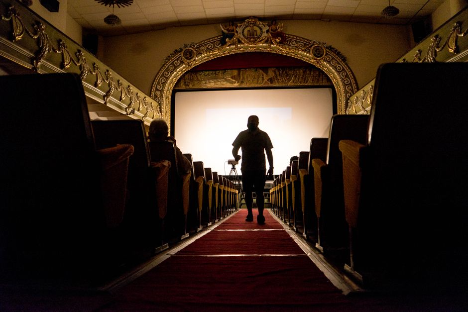 An empty indoor auditorium featuring rows of upholstered black chairs arranged in front of a stage with a large white projection screen. The stage area has a wooden floor and is illuminated by overhea