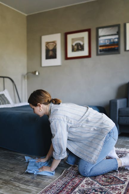 A woman with brown hair tied back, wearing a light blue striped shirt and jeans, is kneeling on a patterned area rug while cleaning a dark blue upholstered bed frame with a cloth in a well-lit bedroom
