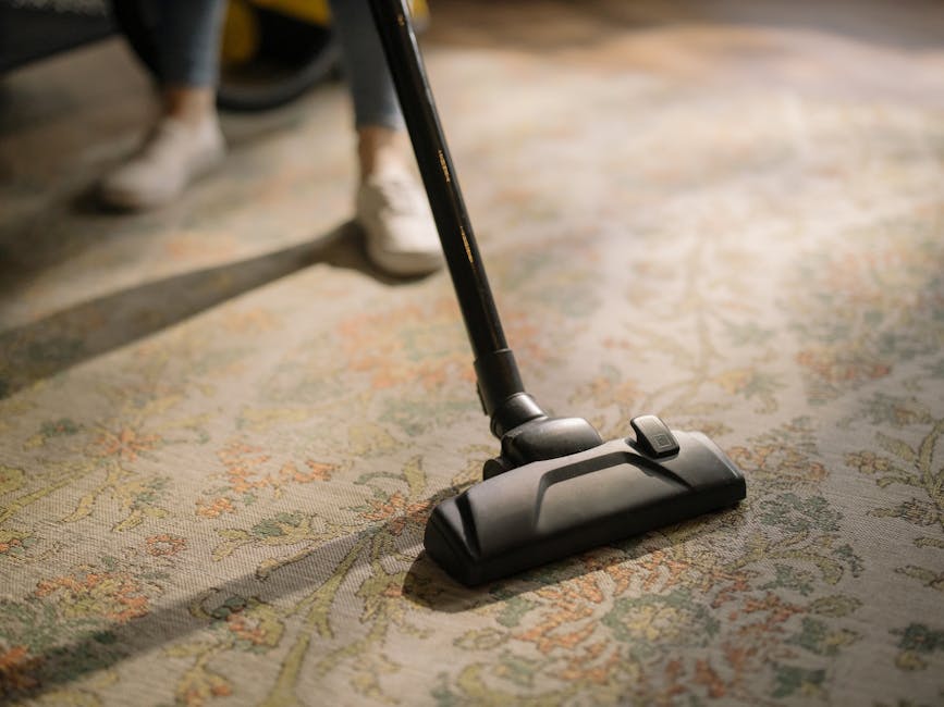 A woman with brown hair tied back, wearing a light blue striped shirt and jeans, is kneeling on a patterned area rug while cleaning a dark blue upholstered bed frame with a cloth in a well-lit bedroom